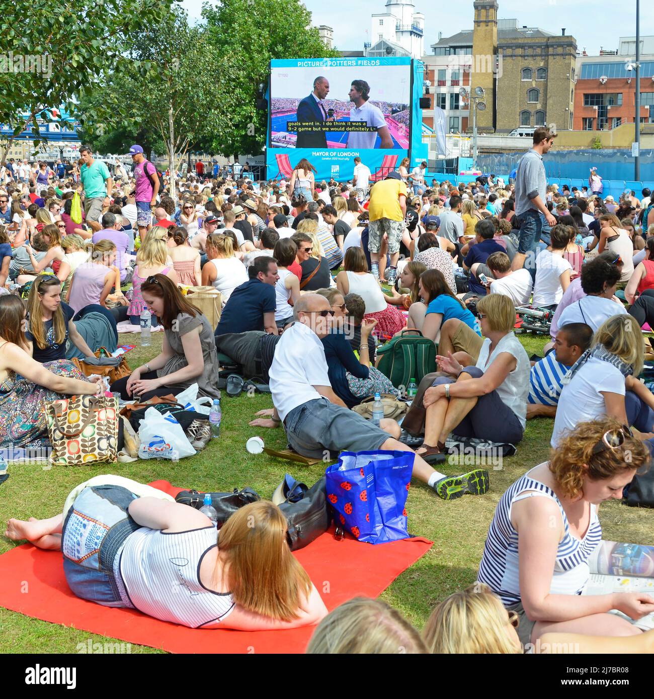 Crowd of people sit on grass watching London 2012 Olympics TV sport event large outdoor television screen in Potters Fields Park Southwark England UK Stock Photo