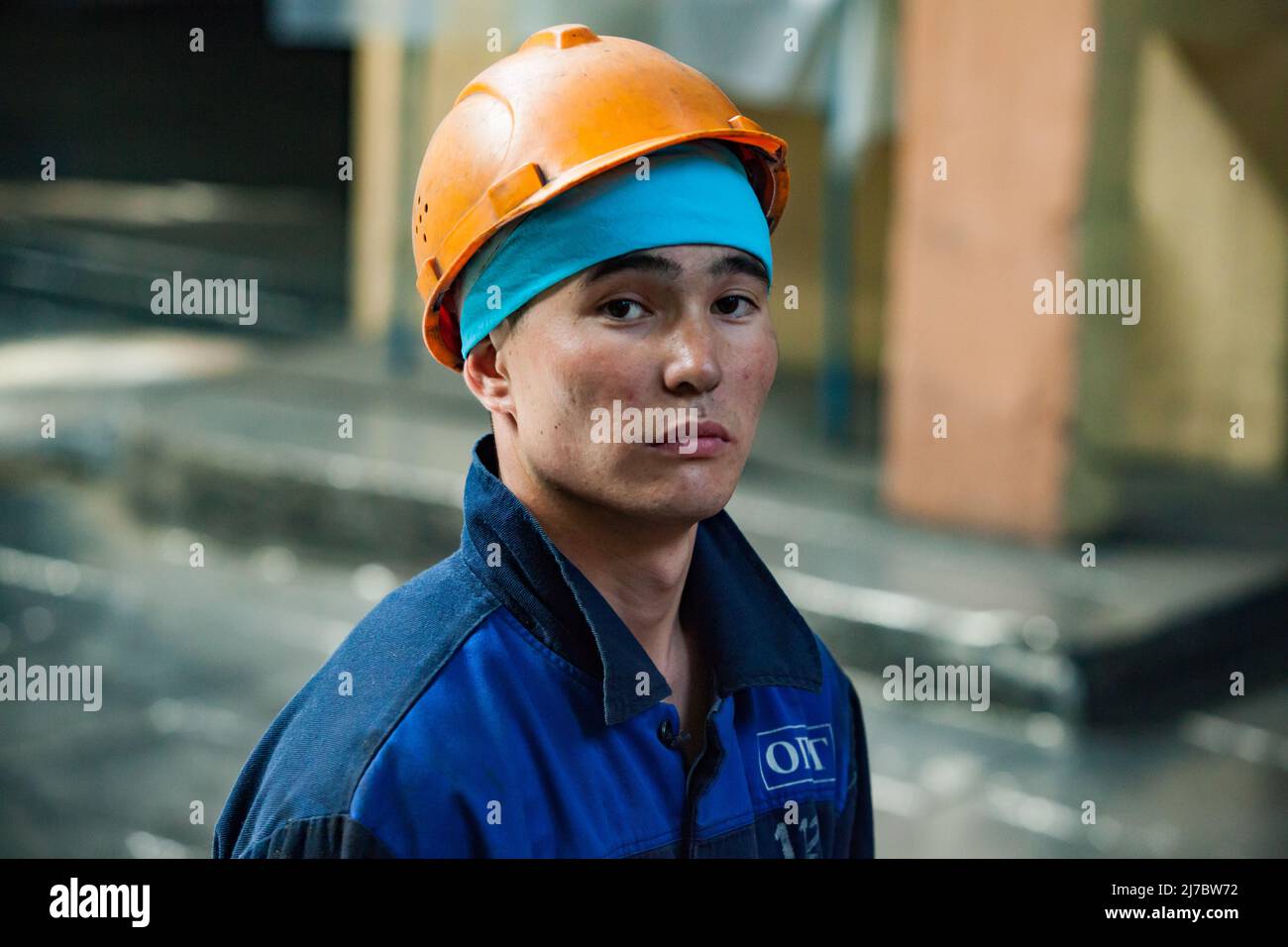Ust'-Kamenogorsk, Kazakhstan - May 31, 2012: Portrait of young Asian worker in blue bandana and orange hardhat. Looks straight in camera. Blurred back Stock Photo