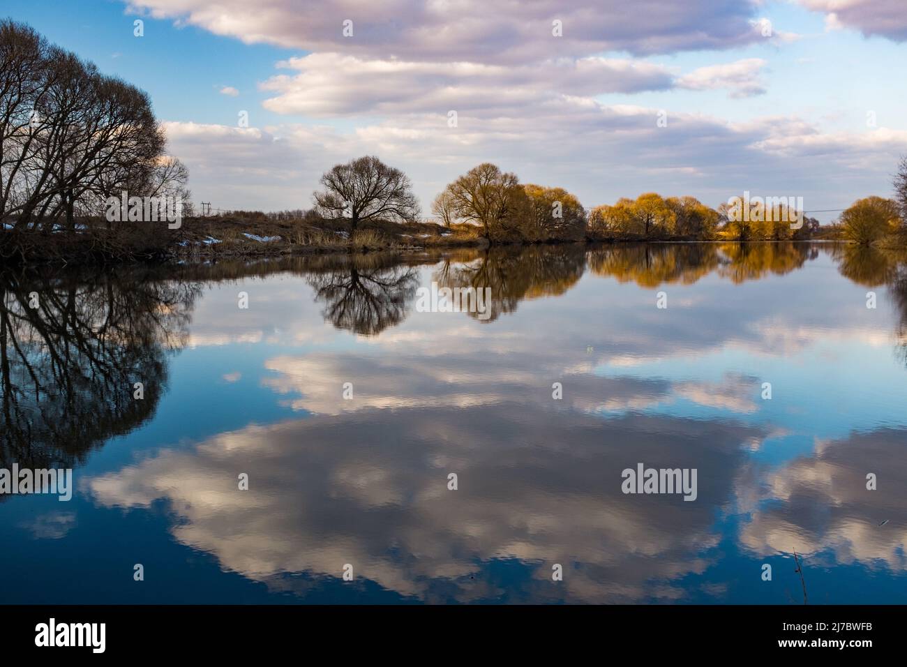 River in spring symmetrical scape. Clear blue water reflection, bare trees  beautiful clouds on blue sky. Sunset yellow light, evening pho Stock Photo