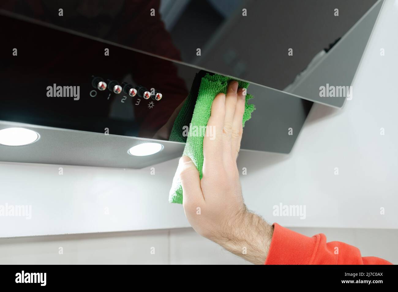 Aspirator cleaning, young man cleans a glossy black extractor hood in a modern kitchen, dusting and wiping Stock Photo