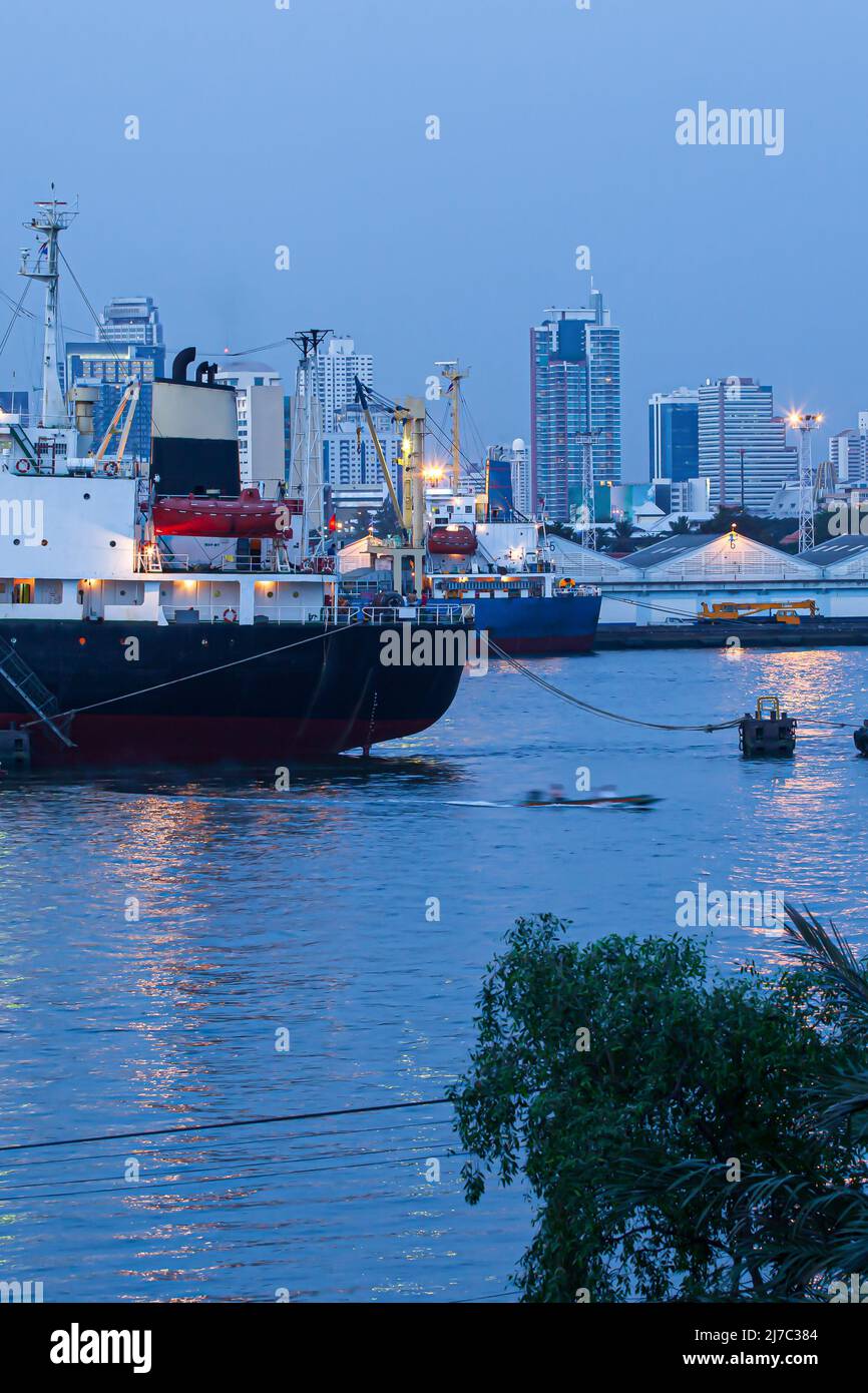 Liners, cargo ships, and container ships dock near the Bangkok Port at dusk, with local boats and other boats passing by. Chao Phraya River, Thailand. Stock Photo