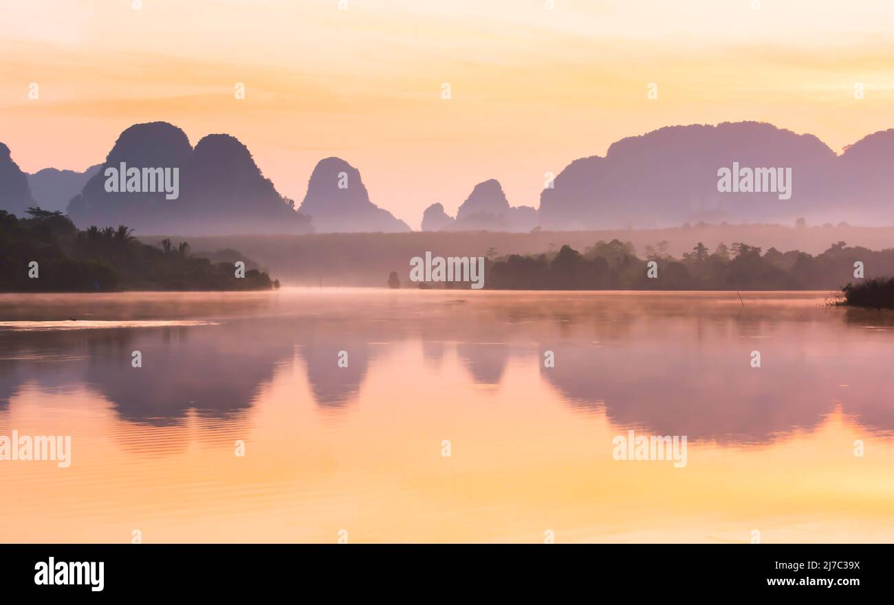 In the landscape of the tropical lake during sunrise, morning mist covers the lake and mountains in the background. Krabi, Thailand. Stock Photo