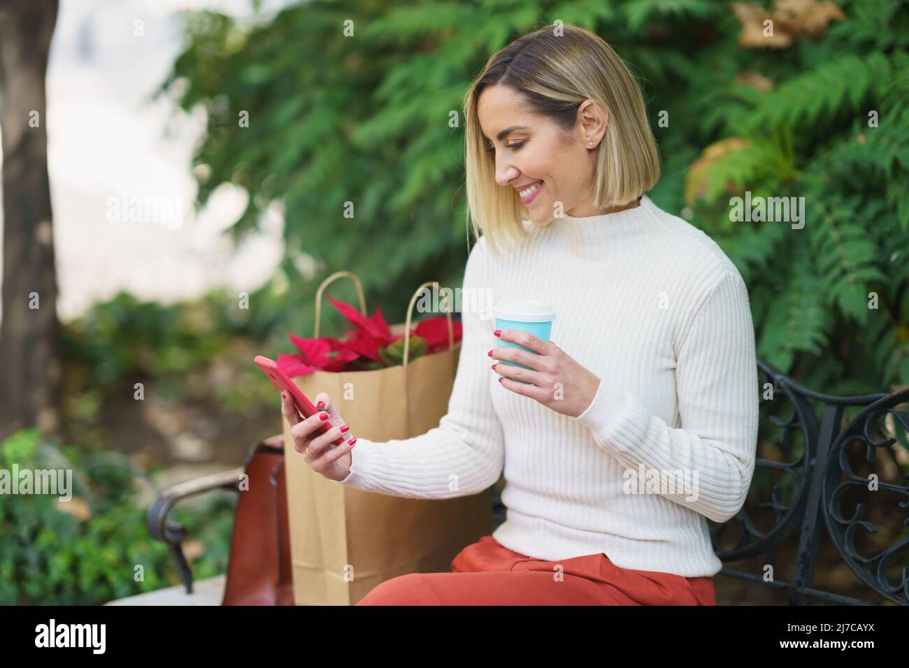 Smiling woman using smartphone and drinking beverage on bench Stock Photo