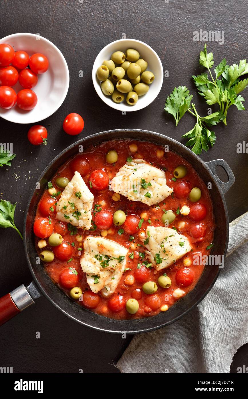 Cod stew with chickpeas, cherry tomatoes and olives in pan over dark ...