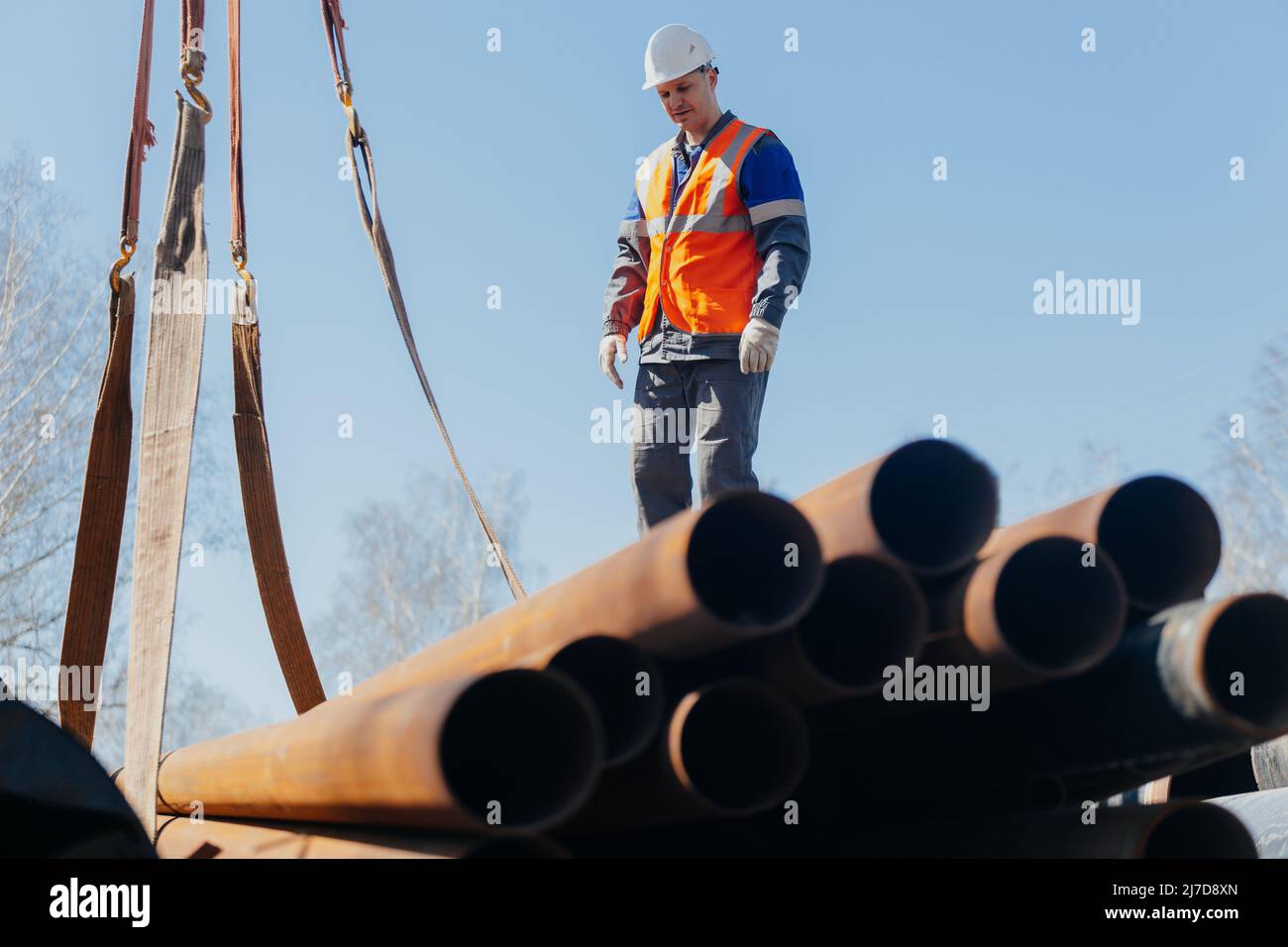Slinger in helmet unloads metal pipes on construction site on sunny summer day. Real scene. Workflow. Real worker. Stock Photo