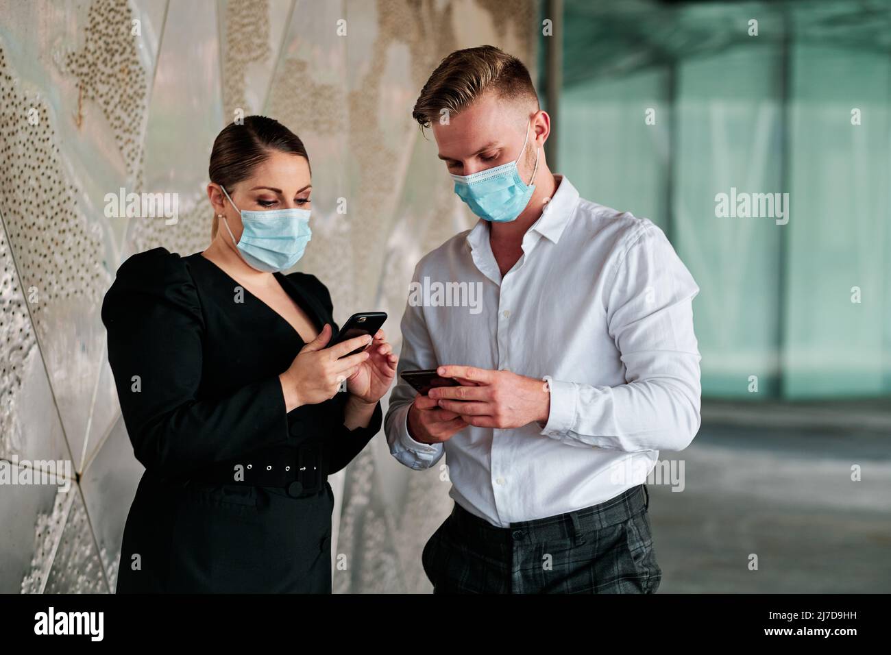 Coworkers with face masks using a mobile phone while talking about business outside the office building. Stock Photo