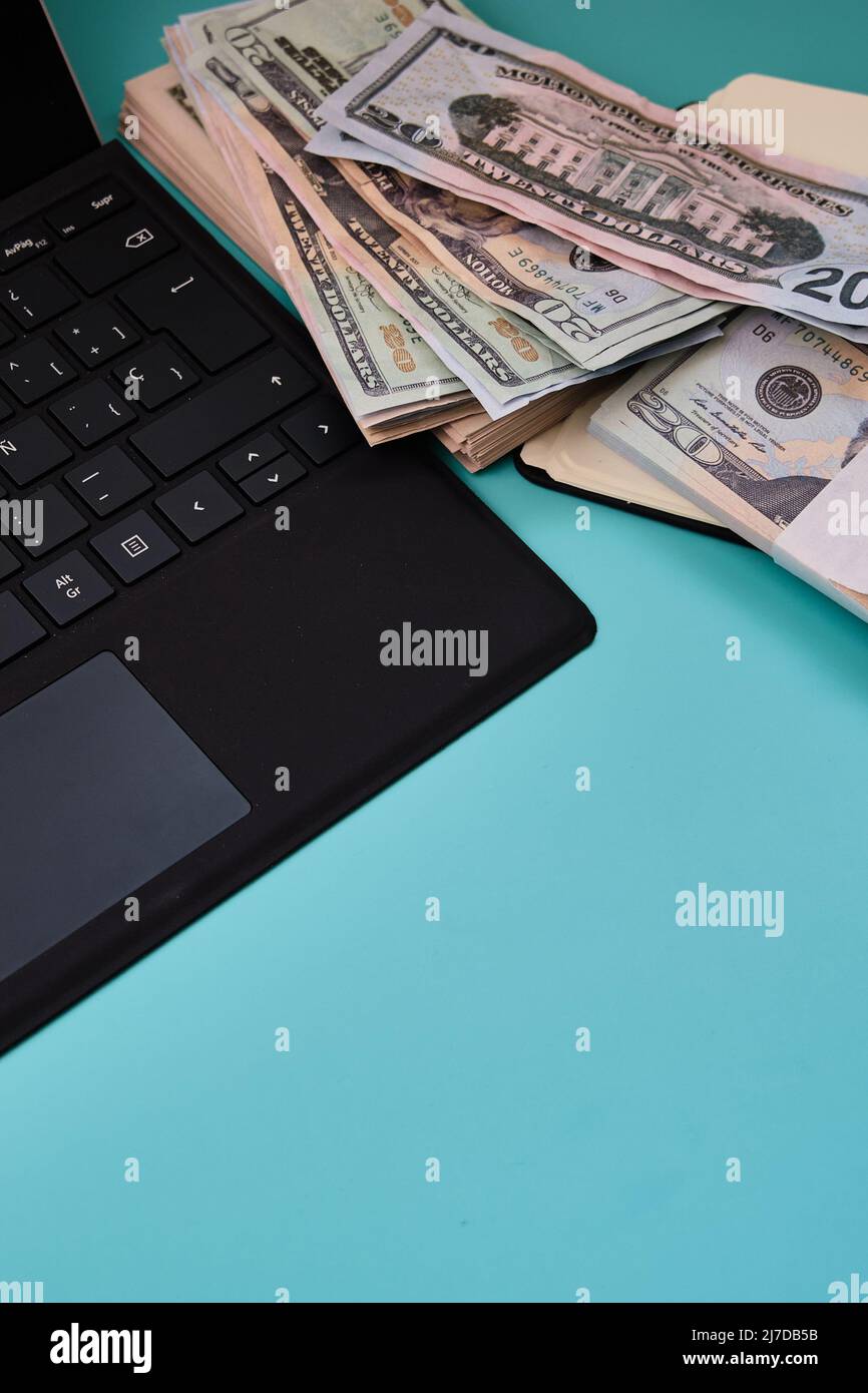 View of a laptop with a pile of dollars next to it, on a light blue table. Photograph in vertical format. Stock Photo