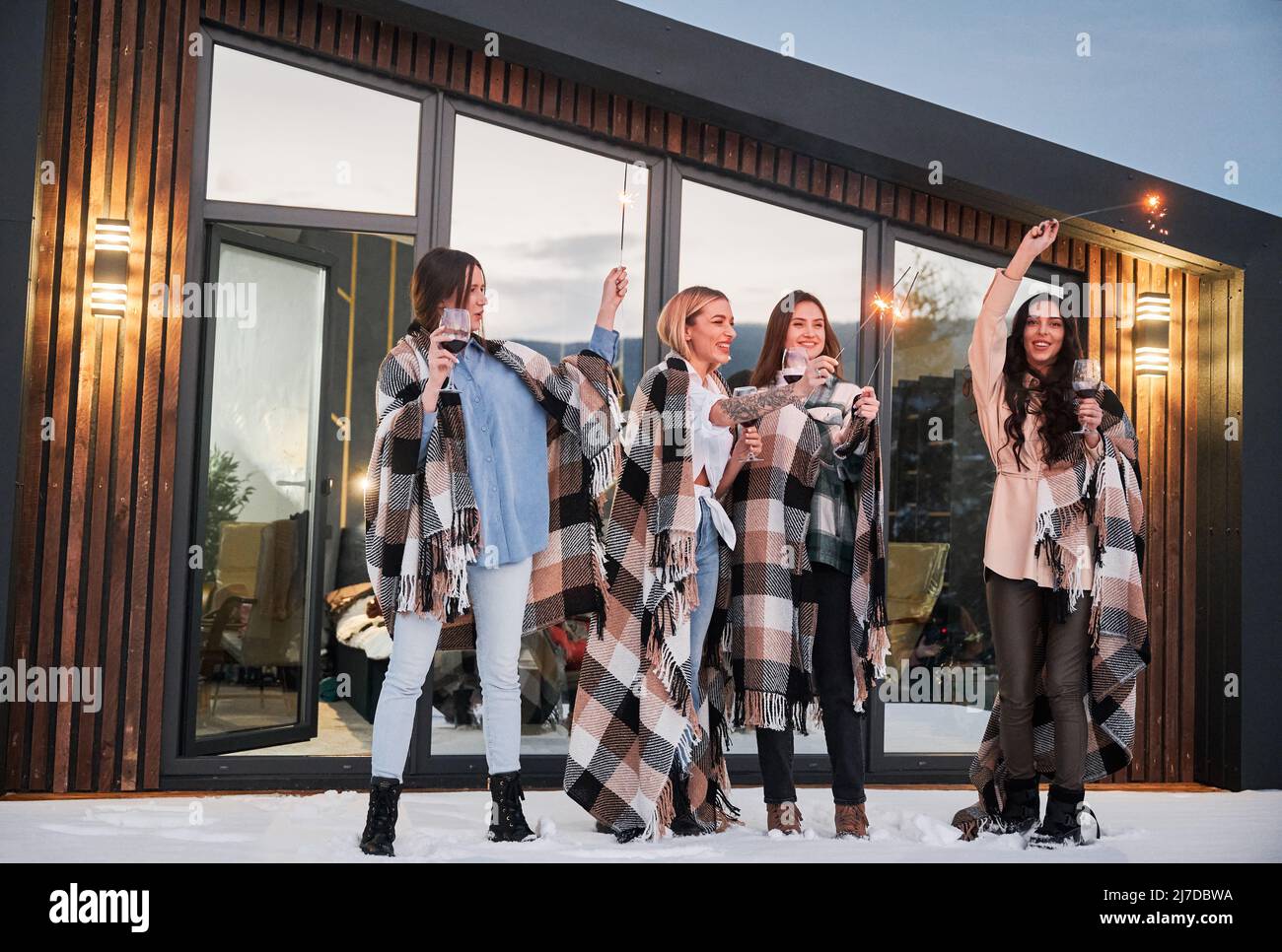 Young women enjoying winter weekends on terrace of contemporary barnhouse. Four girls in plaids drinking wine and celebrating with sparklers in the evening. Stock Photo