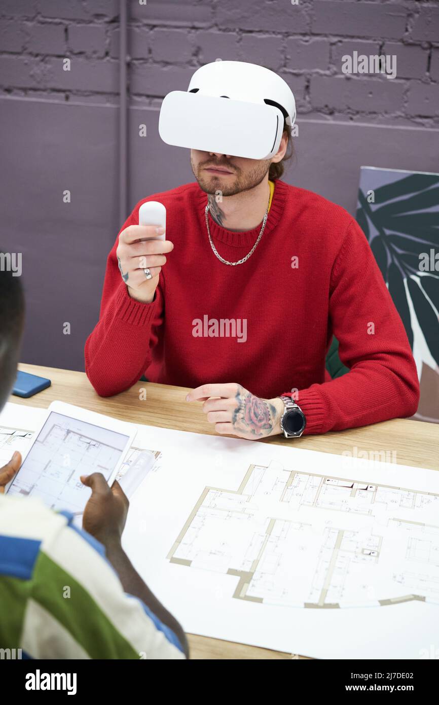 Young designer sitting at table with blueprint and using virtual reality headset to look at new project Stock Photo