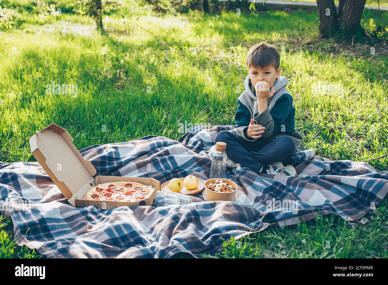 Boy 7-8 years old sitting on plaid and eating apple. Concept of spring or summer picnic Stock Photo