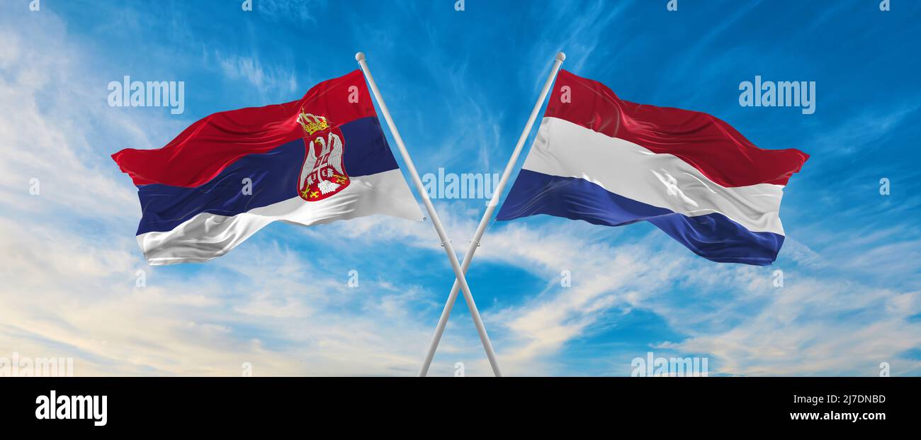 two crossed flags Serbia and Netherland waving in wind at cloudy sky ...