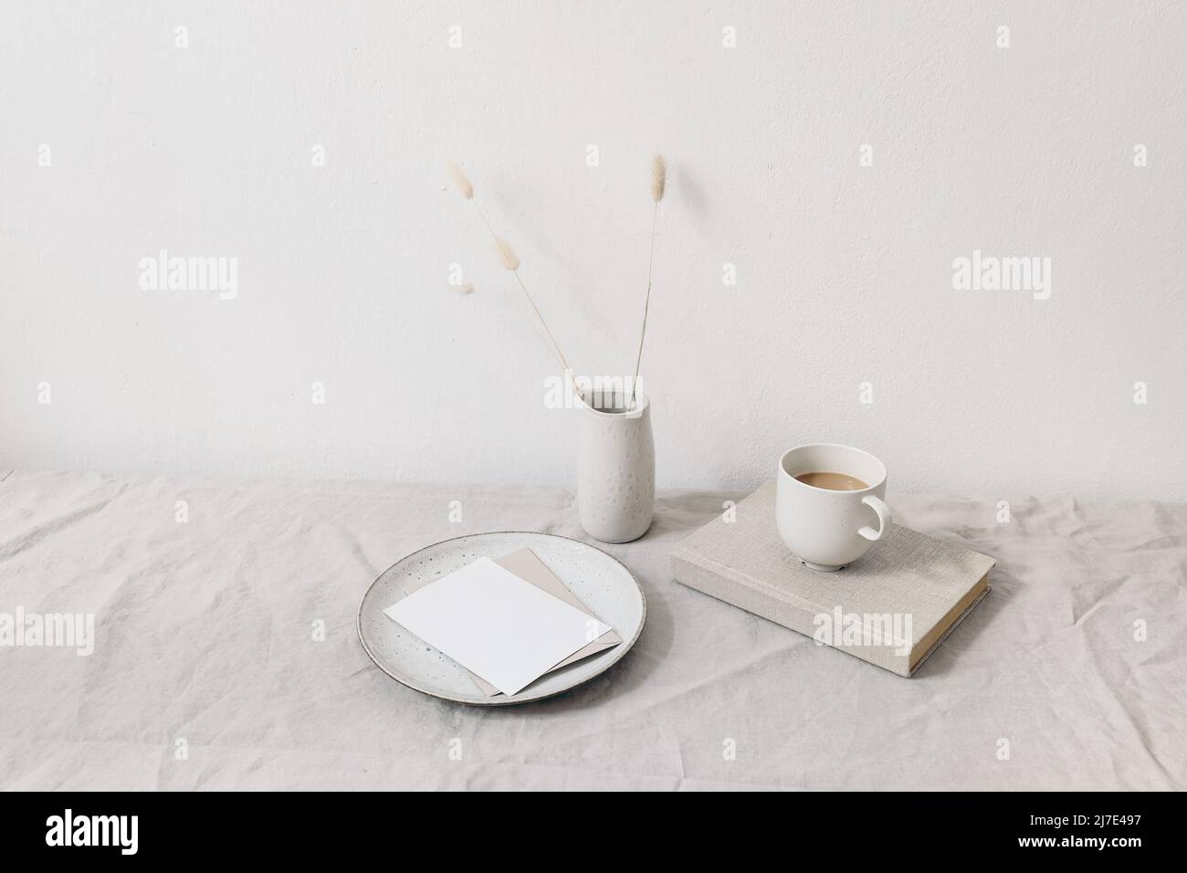 Neutral breakfast still life scene. Blank greeting card, ceramic plate. Vase with dry lagurus bunny tail grass on book. Cup of coffee on book. Linen Stock Photo