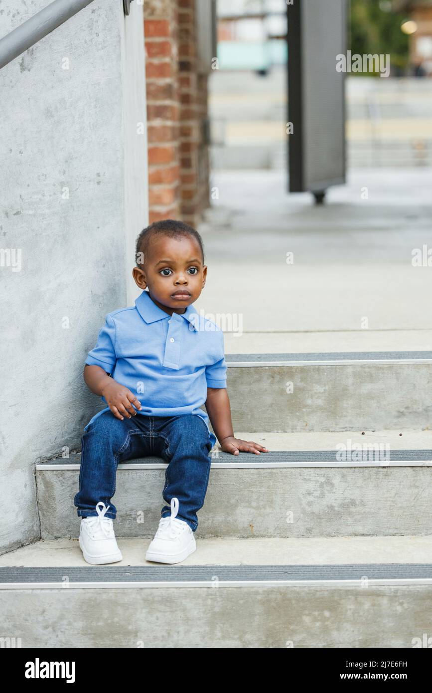 A cute one year old toddler almost preschool age African-American boy with big eyes sitting on city steps Stock Photo