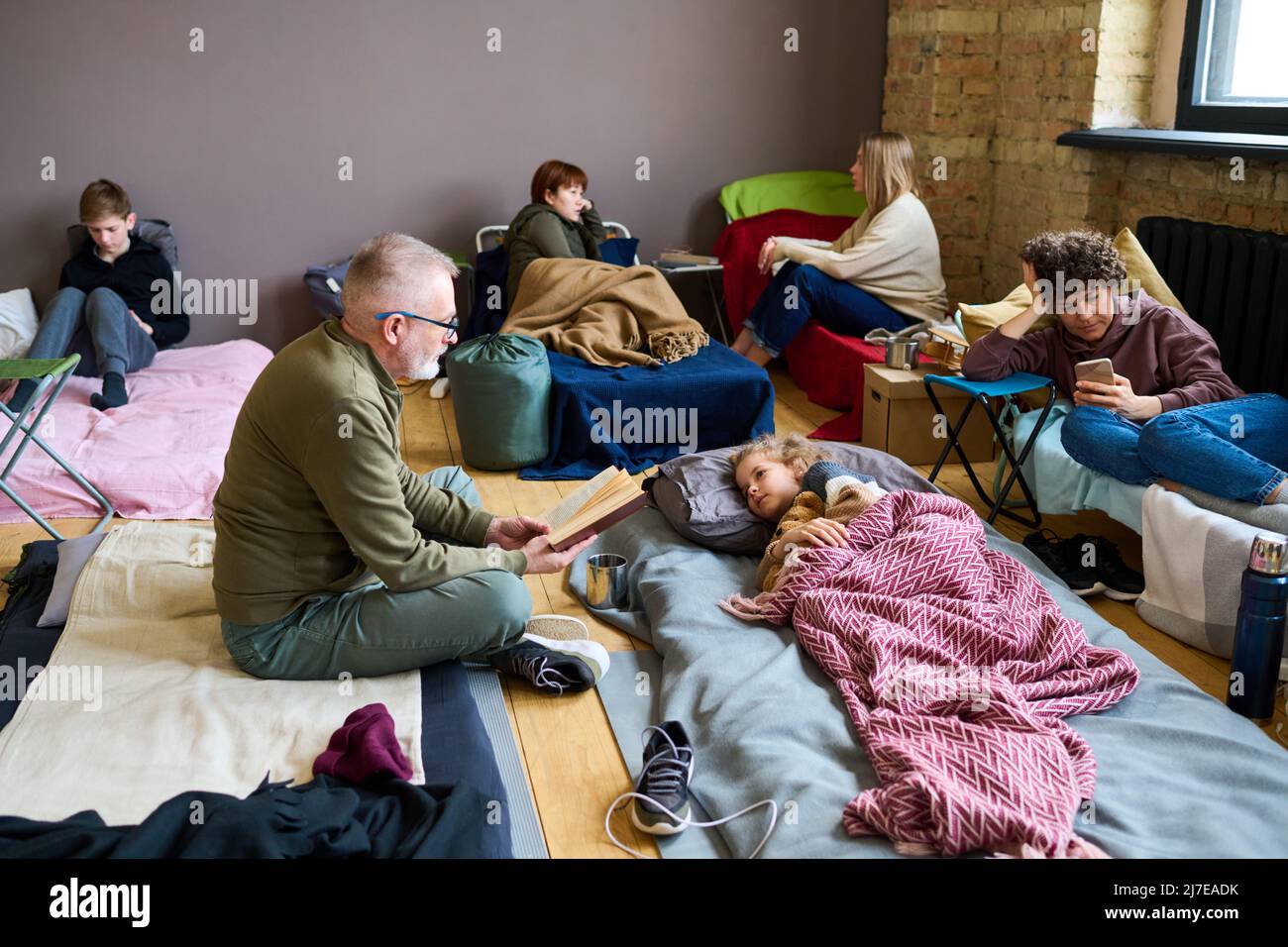 Mature man with open book reading fairy tale to his grandson lying in bed while resting in temporary shelter with other refugees Stock Photo