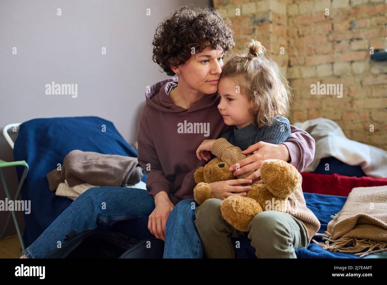 Young woman giving hug to her cute little son with brown soft teddybear while both sitting on sleeping place prepared for refugees Stock Photo