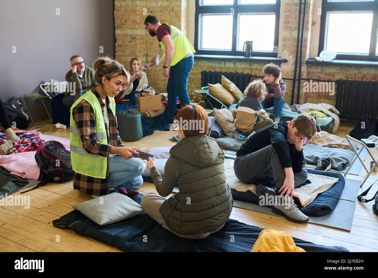 Young female volunteer passing smartphone to one of refugees sitting on mattress on the floor of spacious room serving as camp Stock Photo