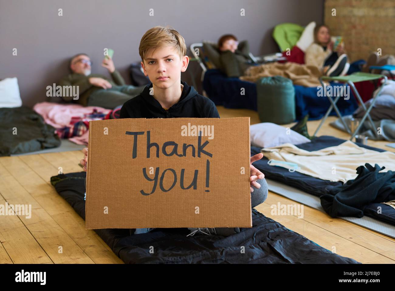 Grateful preteen boy in casualwear holding cardboard poster while sitting on the floor of spacious room serving as temporary shelter Stock Photo