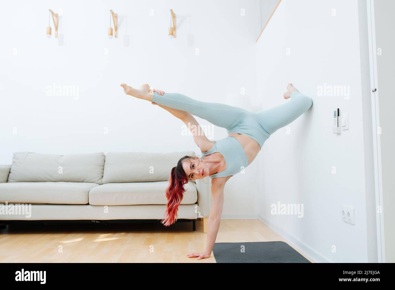 Talanted woman standing on one hand, pressing with knee on the wall at home. Doing beautiful complicated yoga asana on a mat. In a living room. Wearin Stock Photo