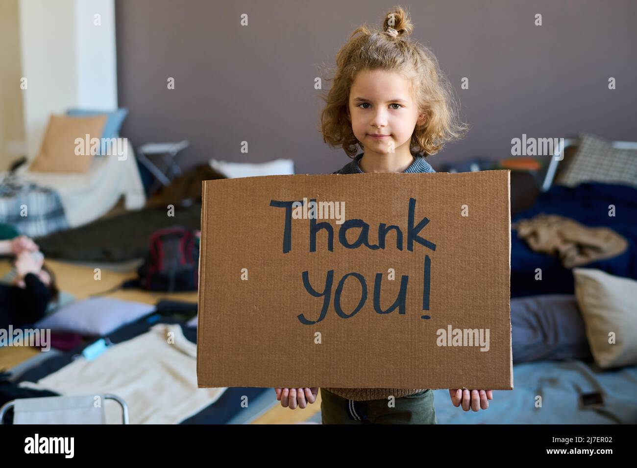 Cute blond little boy with cardboard poster saying thank you looking at camera while standing against sleeping places in refugee camp Stock Photo