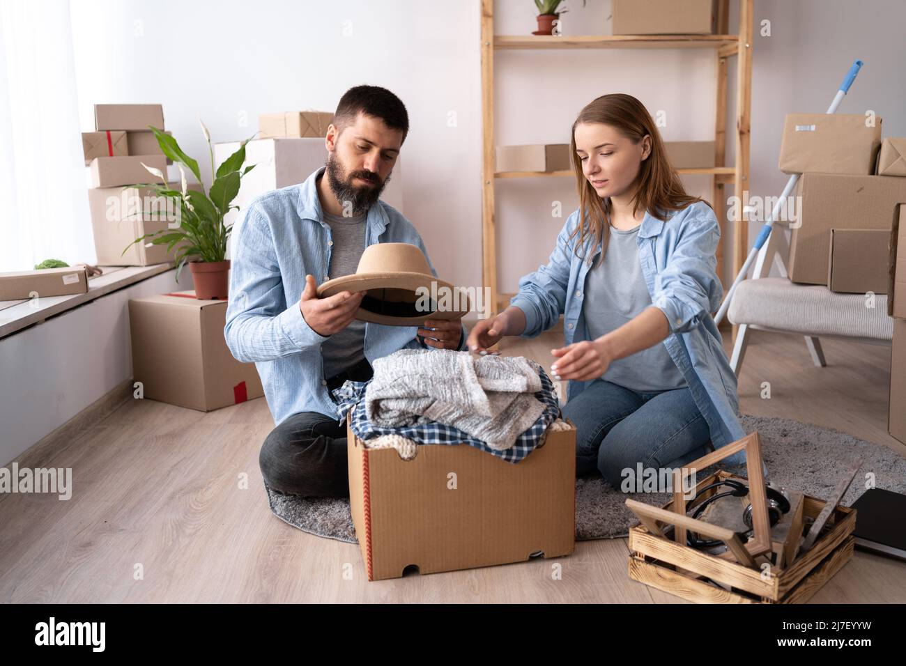a young couple in love in a new apartment on day of the move sit on the floor unpack things a man holds a hat in his hands. The couple moves into a Stock Photo