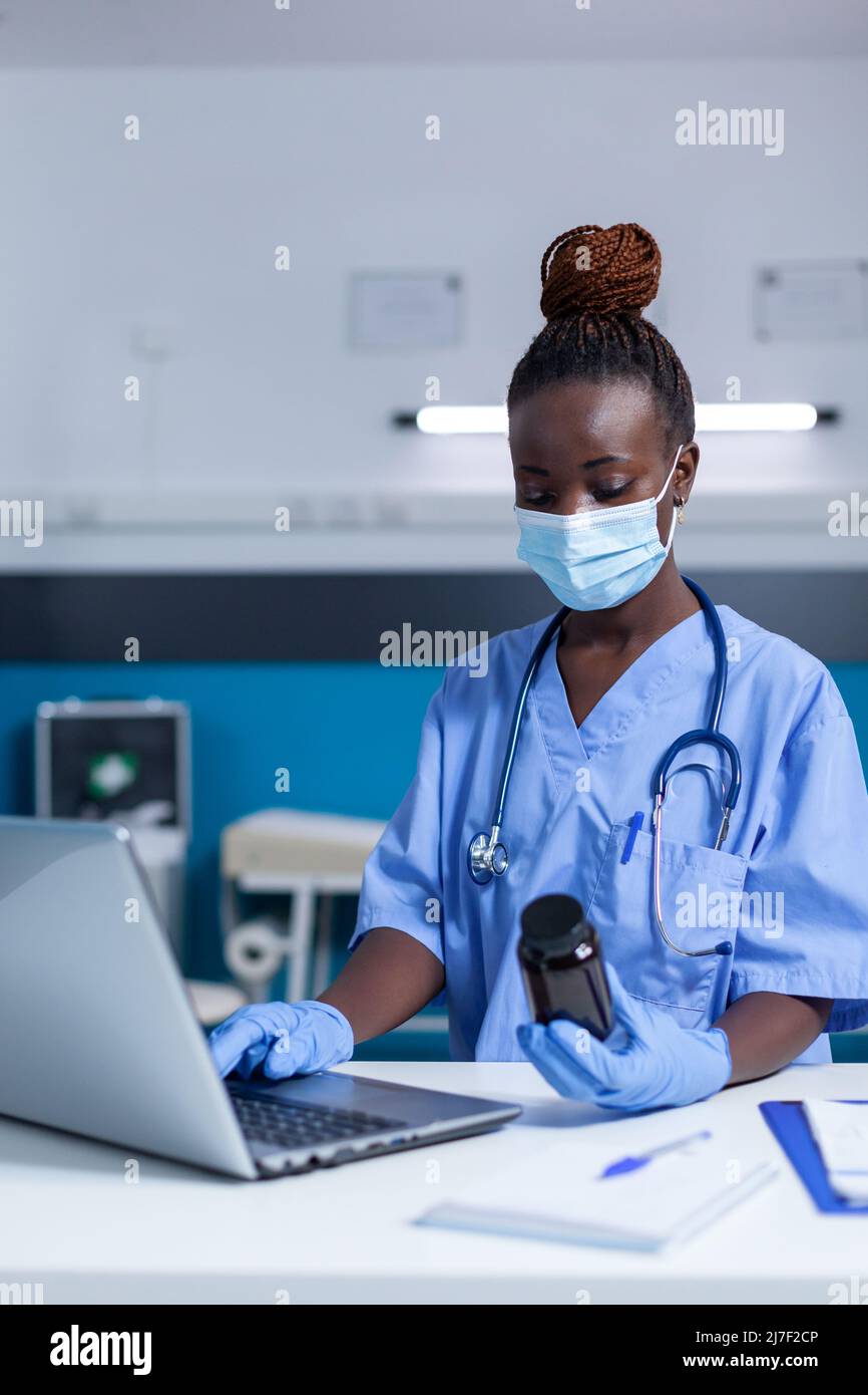 Wearing facemask nurse using modern computer to read prescribed antiviral prospectus. Clinic nurse holding bottle of medicine verifying drug expiry date and ingredients. Stock Photo