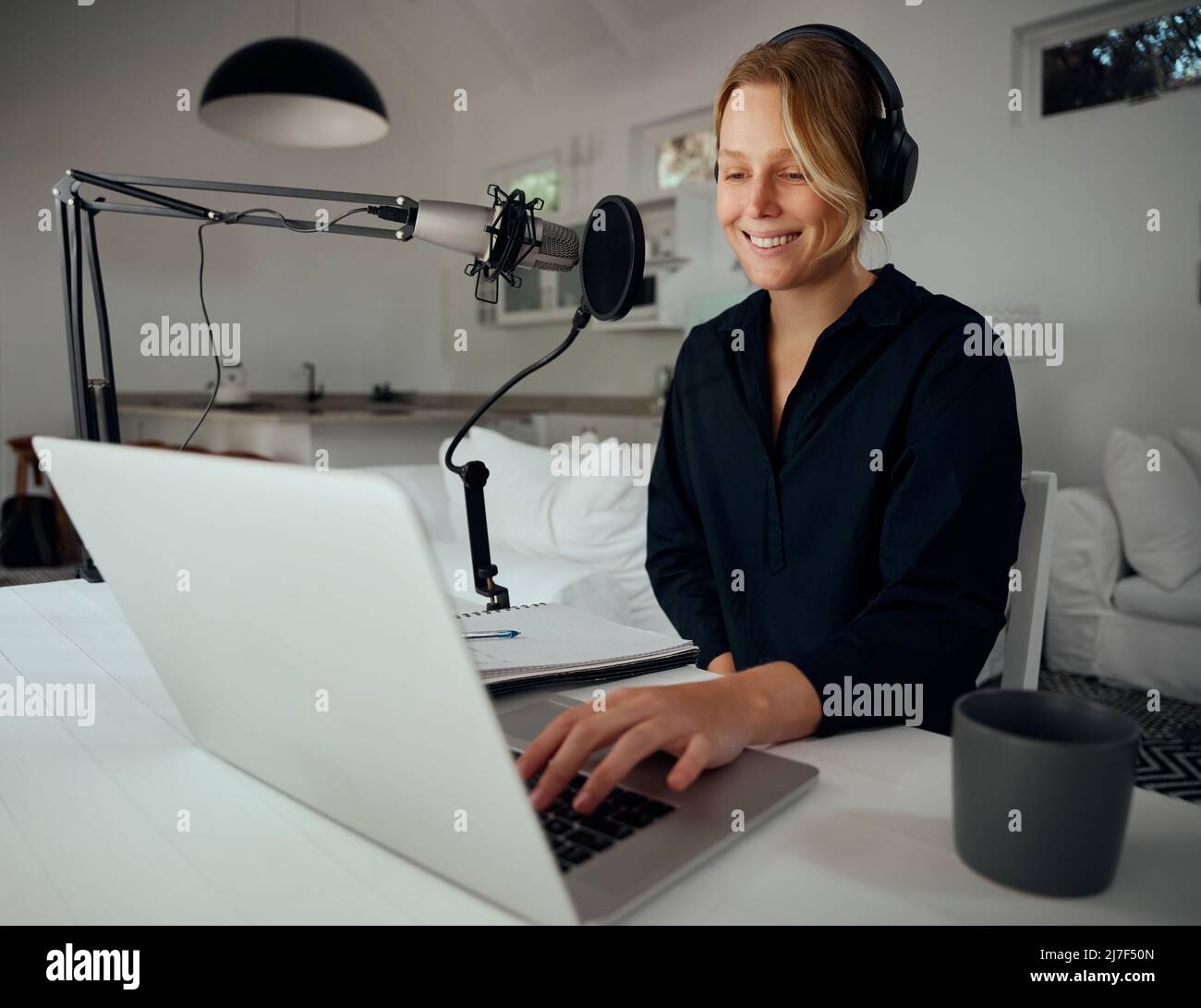 Caucasian female podcaster sitting in home workspace typing on laptop recording podcast  Stock Photo