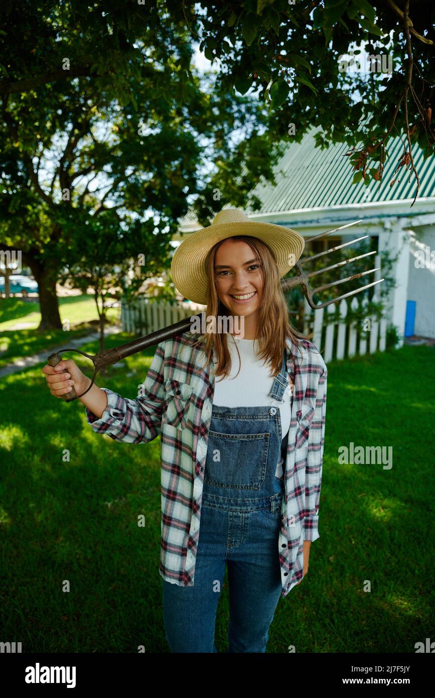 Caucasian smiling female farmer standing by farmhouse holding pitchfork Stock Photo