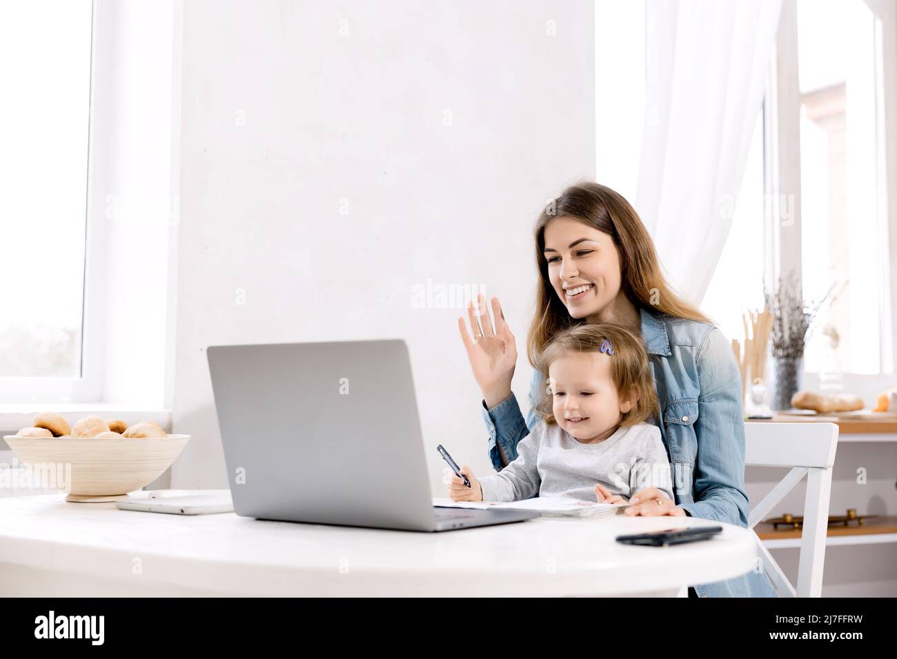 Beautiful mother and little baby works from home, she sits in the kitchen and using laptop for remote work, her cute little girl sits nearby on a Stock Photo