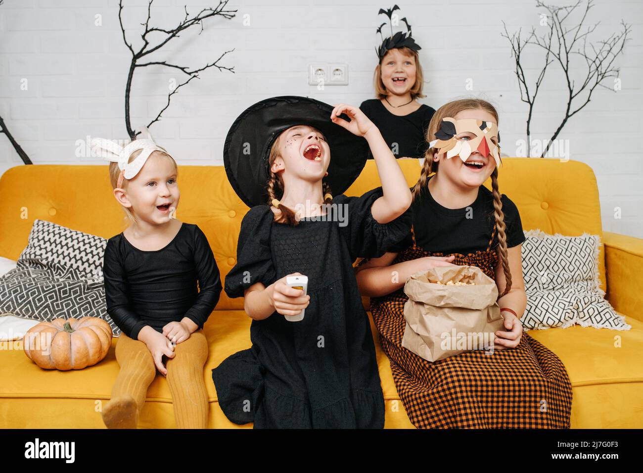 Laughing out loud kids dressed for halloween sitting on a yellow couch, watching funny cartoons. Girl holding remote in her hand. Stock Photo