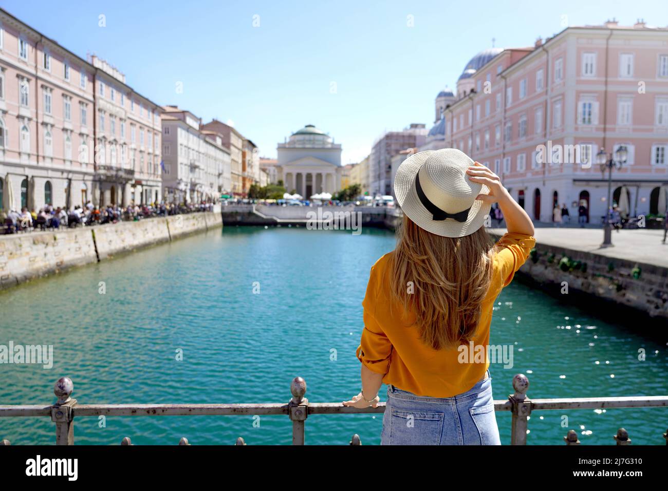 Travel in Trieste, Italy. Back view of pretty girl holding hat looking at Sant Antonio Taumaturgo church on Grand Canal in Trieste, Italy. Beautiful y Stock Photo