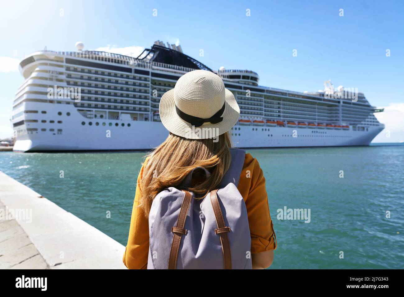 Cruise vacation. Rear view of tourist girl with backpack and hat standing in front of big cruise ship. Stock Photo