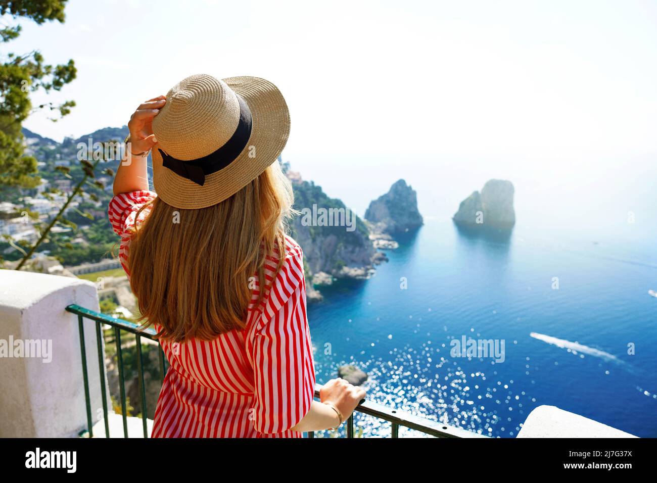 Beautiful young fashion woman with striped dress and hat in Capri Island with Faraglioni sea stack and blue crystalline water on the background, Capri Stock Photo