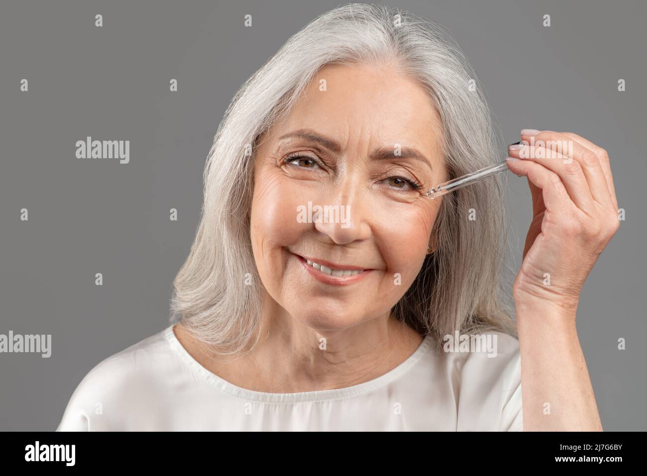 Rejuvenating cosmetics. Beautiful senior woman applying anti-aging serum onto her face over grey studio background Stock Photo