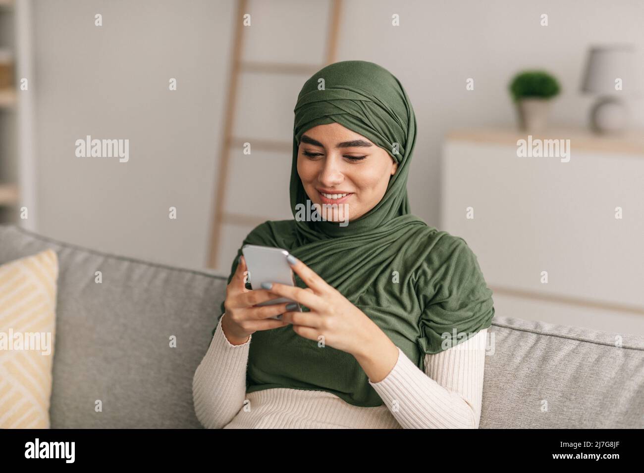 Smiling young Arab female in hijab sitting on couch with cellphone ...