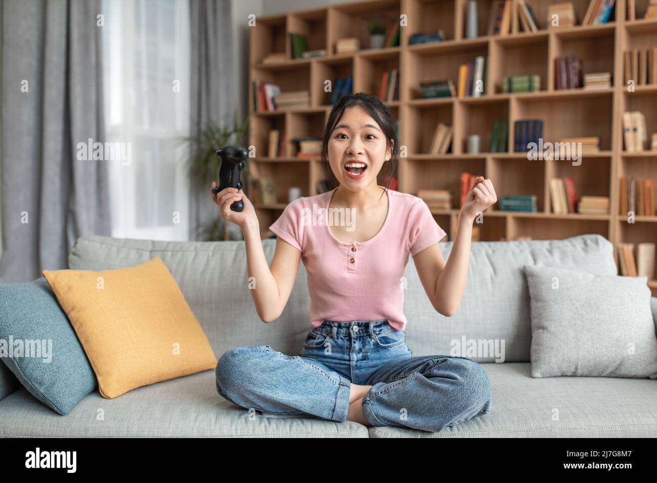 Overjoyed asian female winner playing video games with joystick, sitting on sofa at home, copy space Stock Photo