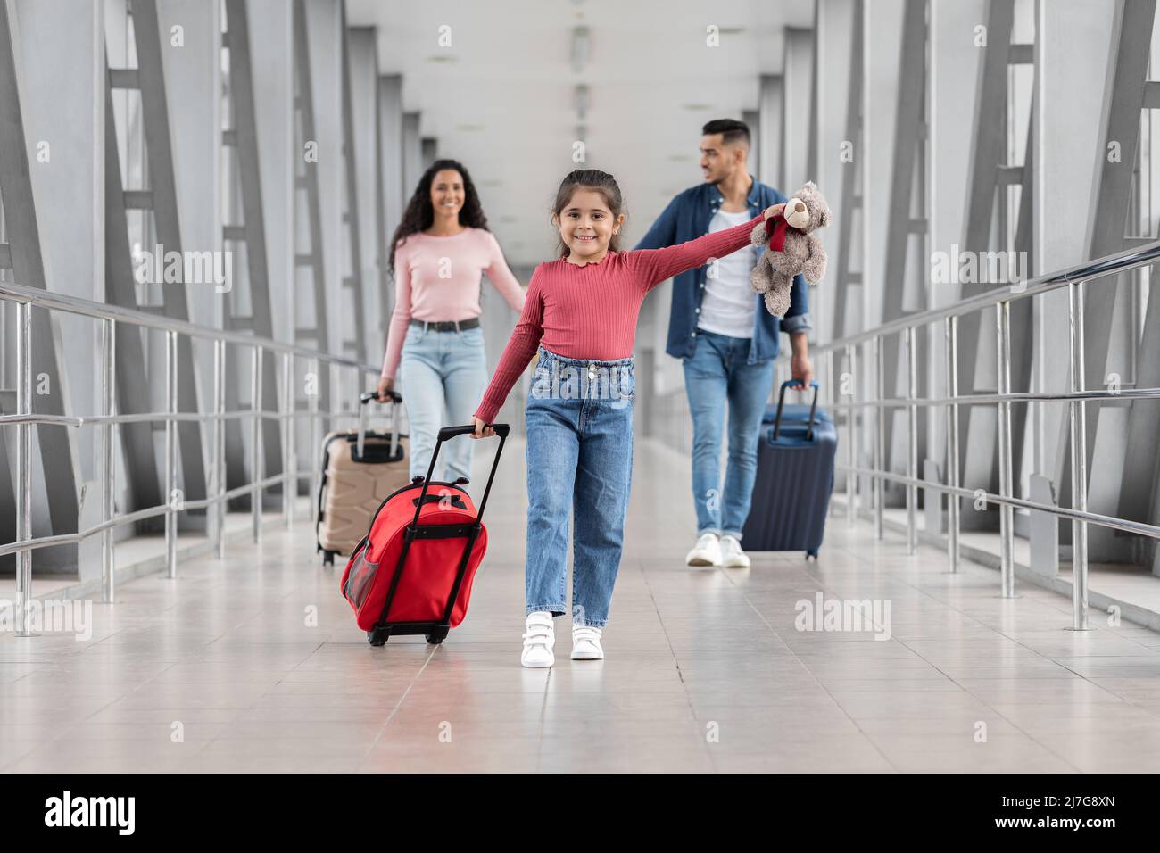 Family Journey. Little Girl Carrying Suitcase While Walking With Parents At Airport Stock Photo