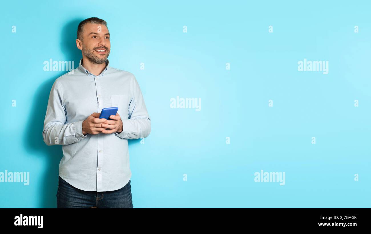Middle Aged Man Holding Cellphone Looking Aside Over Blue Background Stock Photo