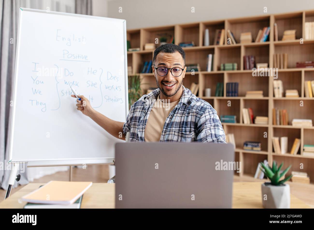 E-learning concept. Male English teacher sitting at table, pointing at whiteboard, having online lesson from home Stock Photo