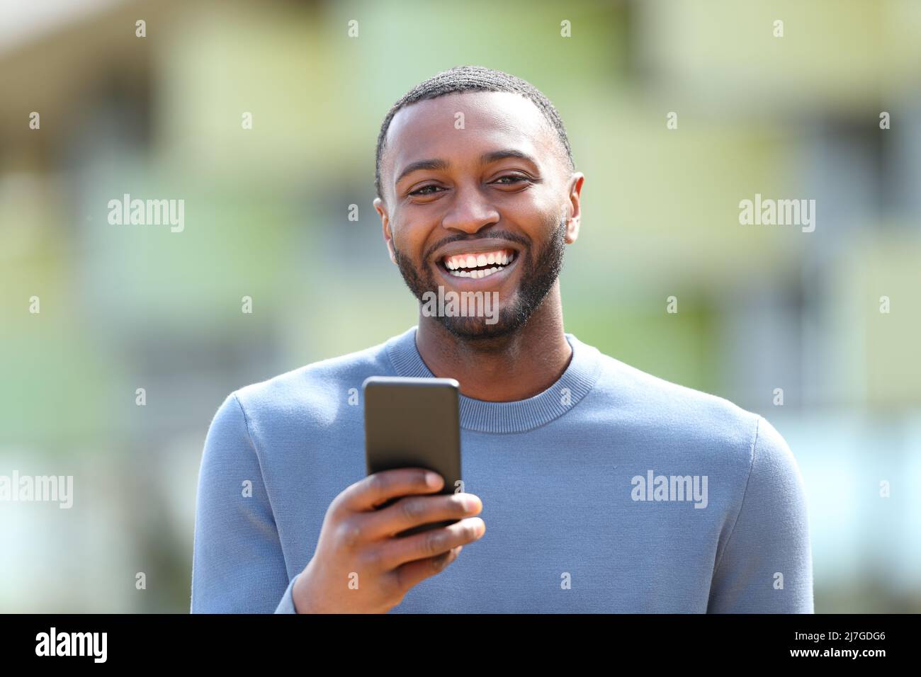 Front view portrait of a happy man with black skin laughing holding phone looking at you in the street Stock Photo