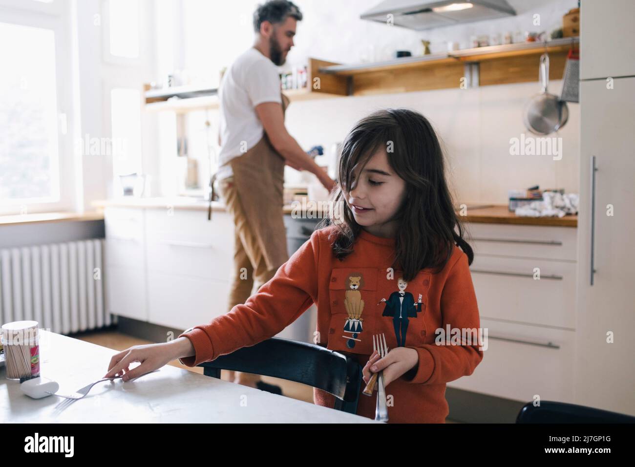 Girl arranging fork on dinging table while father doing chores in kitchen Stock Photo