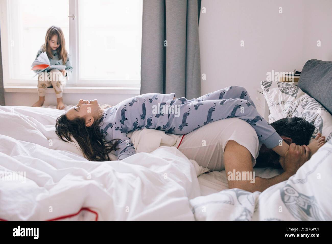 Playful father and daughter lying on bed while girl reading book sitting at window Stock Photo