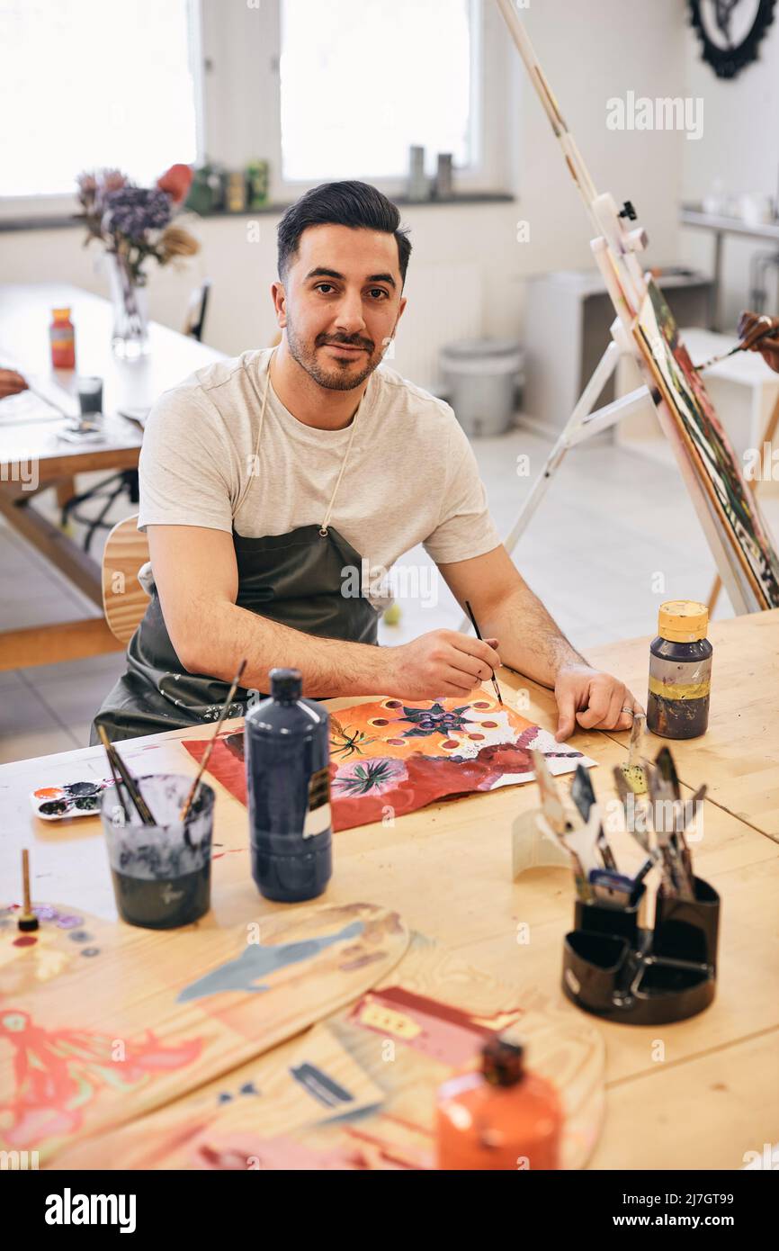 Portrait of confident young male student painting while sitting at table in art class Stock Photo