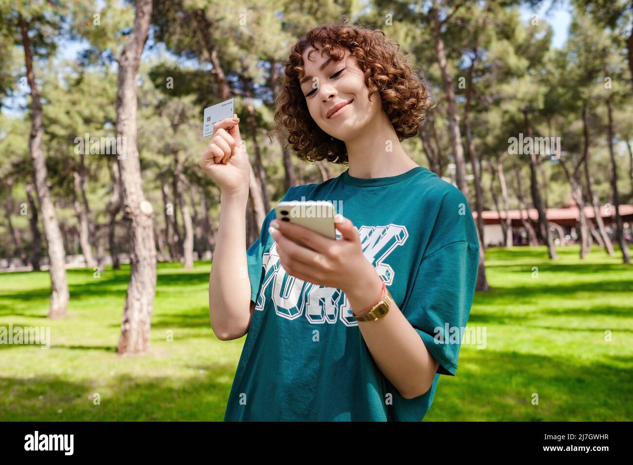 Happy young redhead woman in green tee showing plastic credit card while using mobile phone. Outdoor online or mobile shopping concepts. Stock Photo