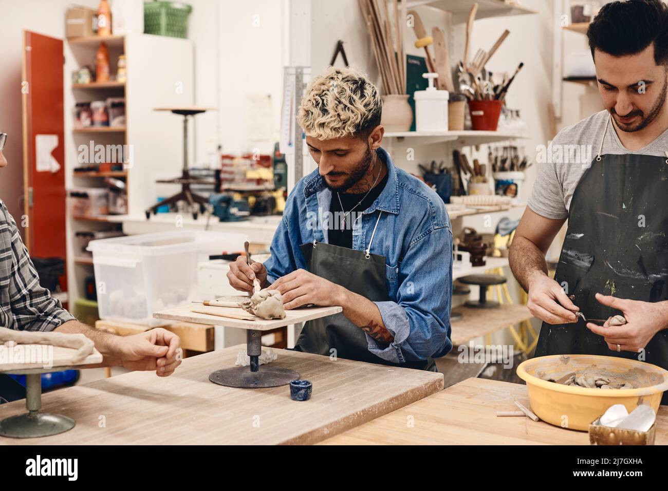 Young man using work tool by friend molding clay while standing at table in art class Stock Photo