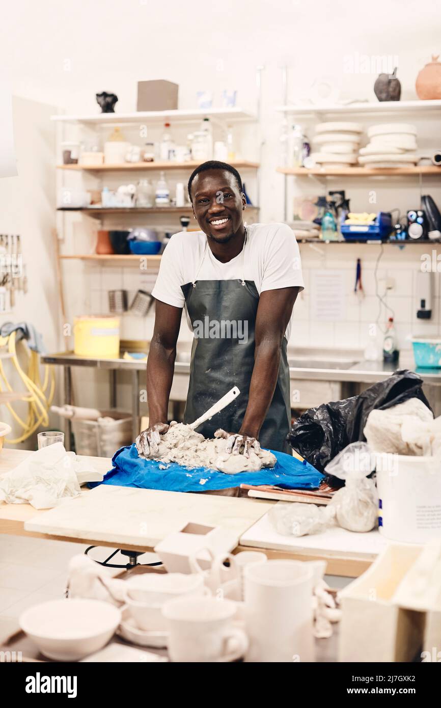 Portrait of happy young artist wearing apron standing with clay on table in art class Stock Photo