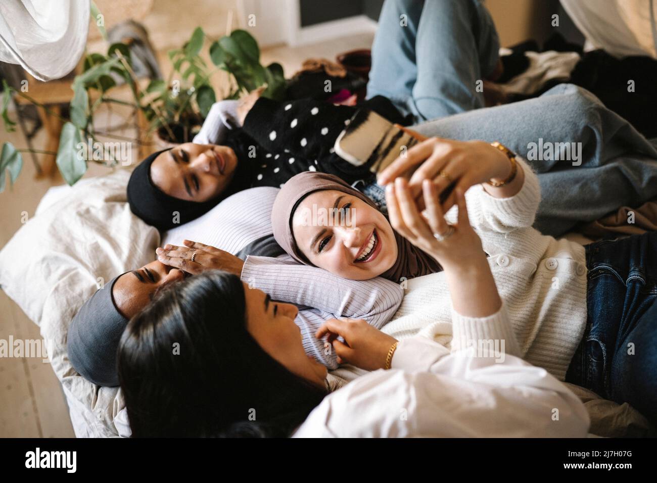 Happy woman wearing hijab taking selfie with friends lying on bed at home Stock Photo