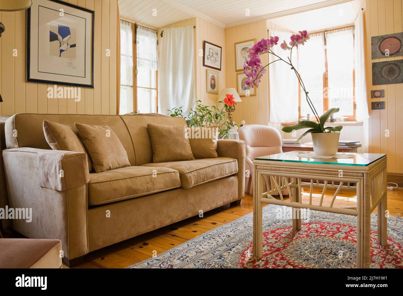 Tan upholstered sofa, armchair, wicker table with glass top and blue and red weaved rug in living room inside old 1746 home. Stock Photo