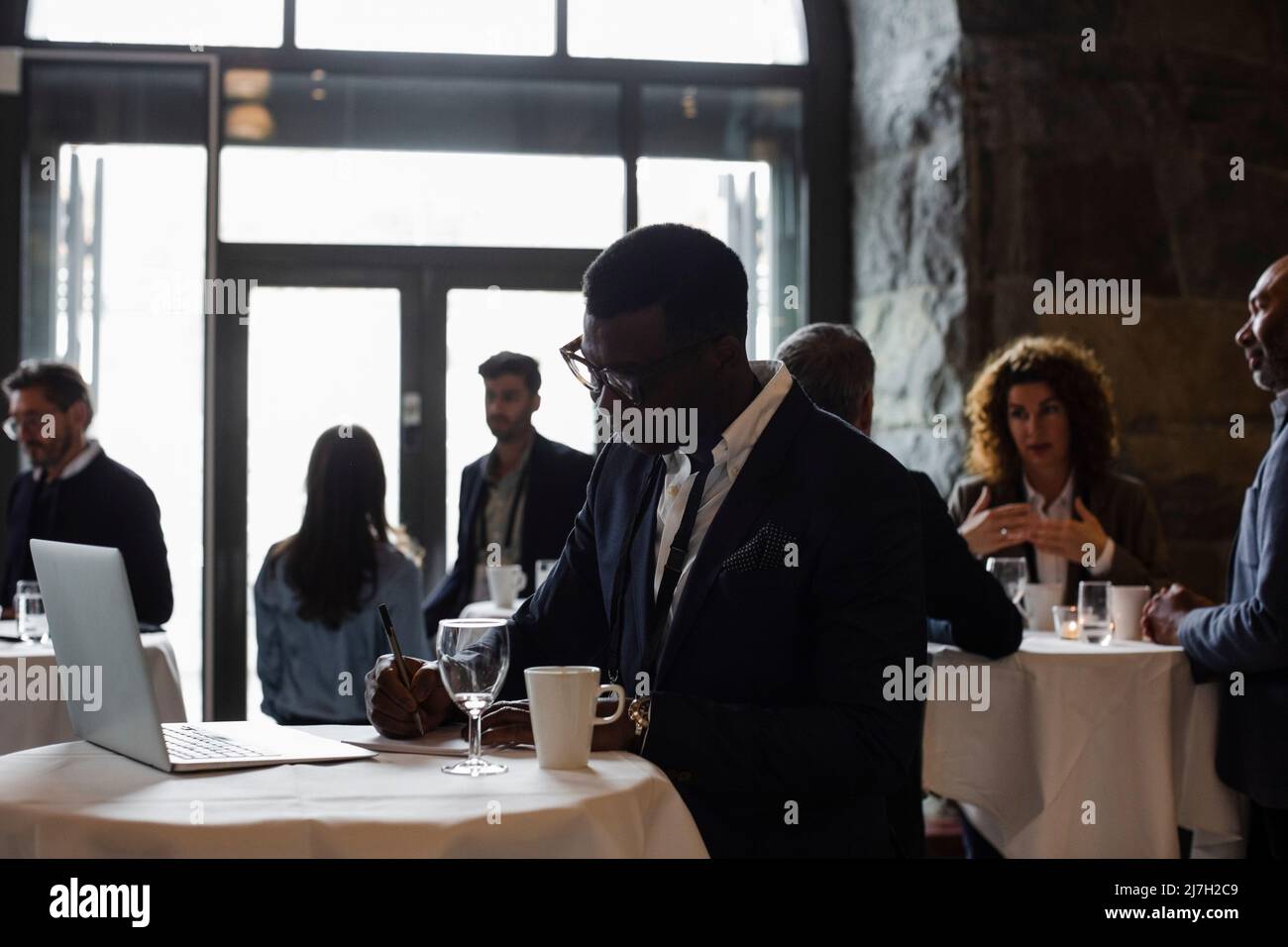 Businessman with laptop on table writing in notepad while standing at seminar Stock Photo