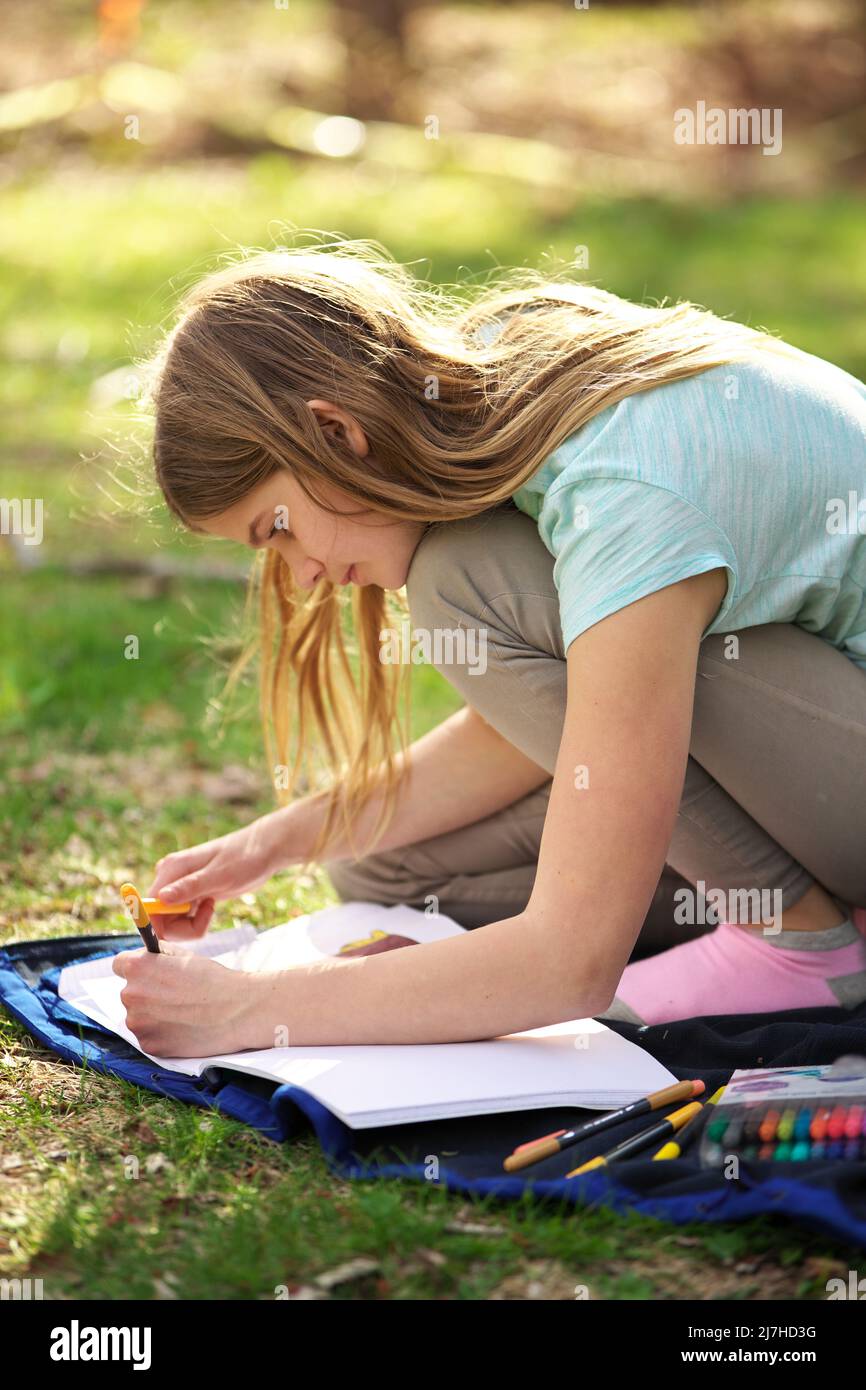 A young adolescent girl writes or colors in a notebook or journal on a blanket on the grass  Stock Photo