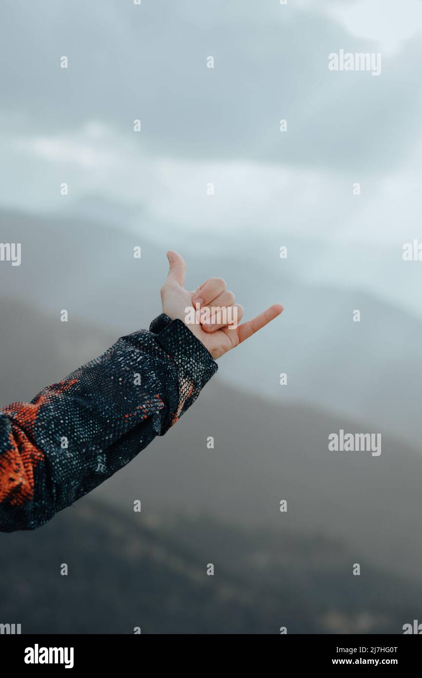 Unrecognizable male hand wearing a coat making the hang loose sign on top of a mountain with peaks in the background Stock Photo